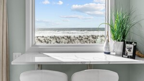 Ein Zimmer mit großem Fenster und Meerblick, einem weißen Schreibtisch, zwei Stühlen, einer Topfpflanze und einem gerahmten Foto.