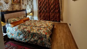 Ein Schlafzimmer mit floralem Bettüberwurf, einem roten Teppich mit Muster und Holzboden, sowie einer Deckenleuchte.