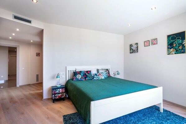 A bedroom with a white bed, green bedding, a blue rug, and a nightstand with a lamp.