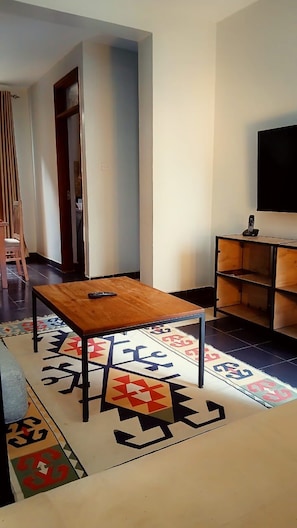 Una sala de estar con una mesa de madera, un tapete estampado, un televisor y una estantería.