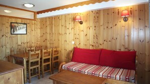 Een knus kamer in chaletstijl met houten wanden, een houten eettafel met stoelen, een rode bank met decoratieve kussens en twee wandlampen.