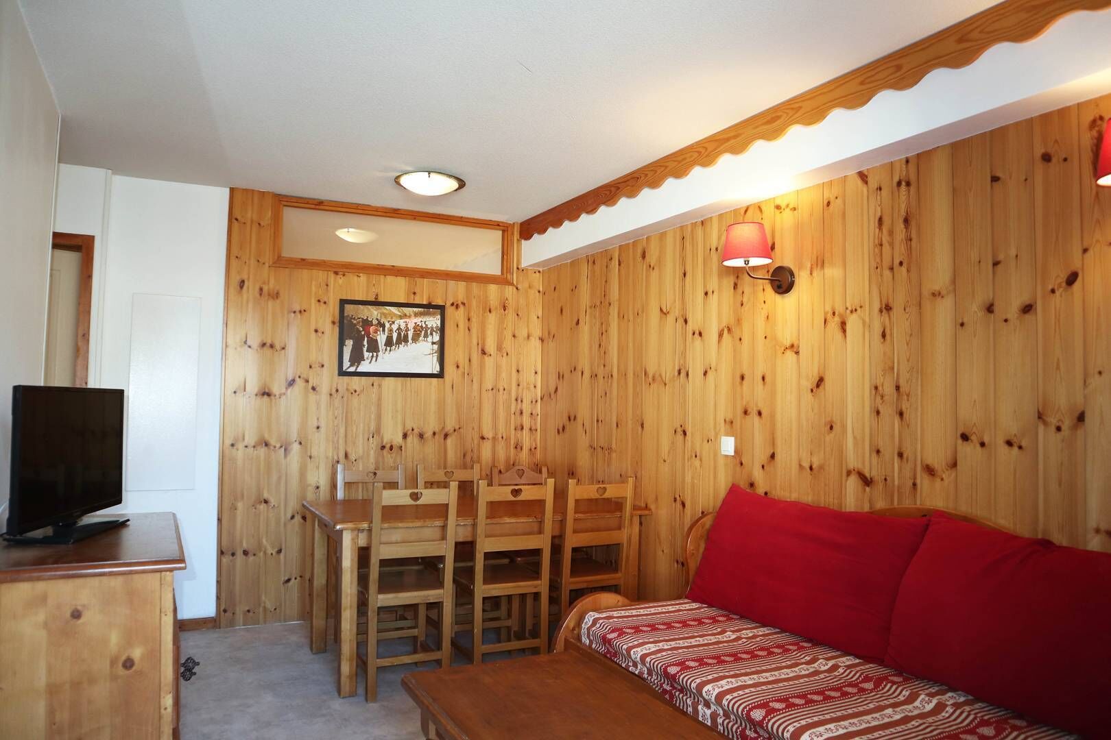 A cozy room with wooden walls, a dining table with chairs, a red sofa, and a television on a wooden cabinet.