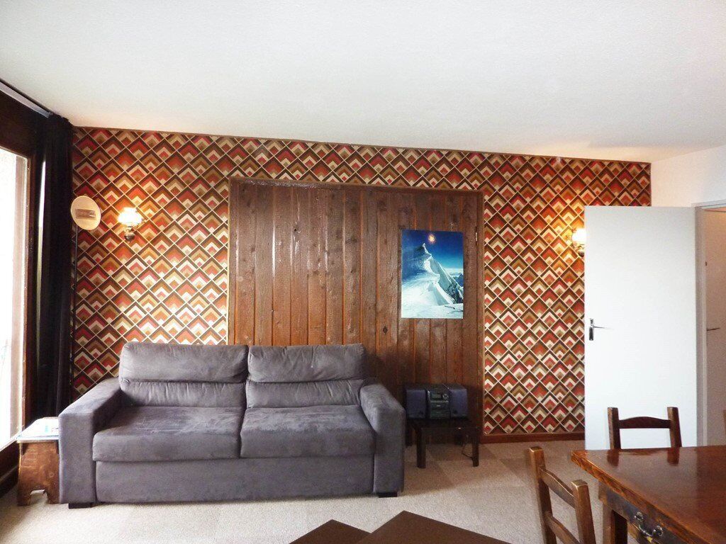 A living room with a grey sofa, wooden panel wall, patterned wallpaper, a painting, and a dining area with a wooden table and chairs.