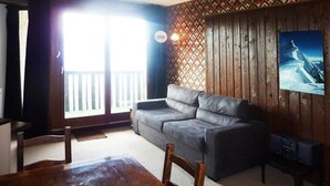 Un salón con sofá gris, paredes de madera, una mesa de comedor de madera y un ventanal con vistas a un balcón.