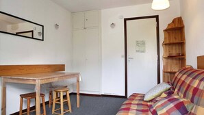 A small, cozy room with a wooden desk, two stools, a sofa with a colorful blanket, a mirror, a wardrobe, and a wooden shelf with a basket. - 32 LES Gradins lES Orres 1650 (LES ORRES 1650)