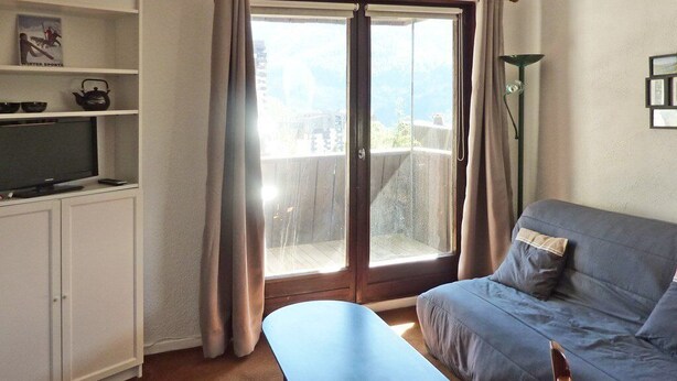 A living room with a sofa, a bench, a TV, and a sliding glass door to a balcony. - 119 LE Sunny Snow 1650 (LES ORRES 1650)