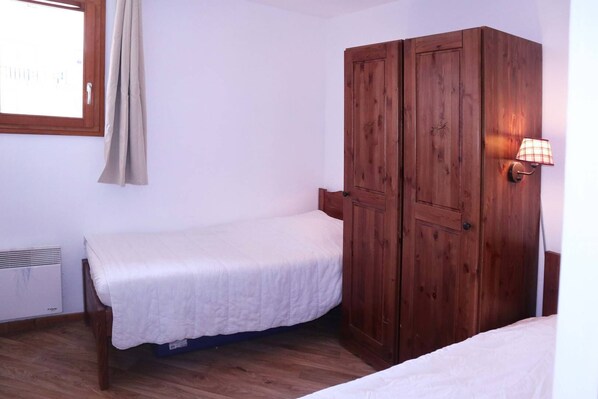 Un dormitorio pequeño con una cama individual, un armario de madera, una ventana con cortinas y una lámpara fijada a la pared.