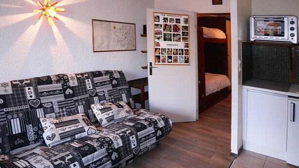 A cozy room with a patterned sofa, a map on the wall, a door with a 'Nos Merveilleux' sign, and a kitchenette with a microwave.