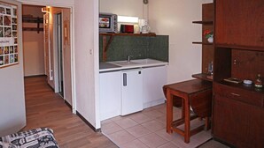 A compact kitchen with a microwave, coffee maker, and a small sink. There is a wooden table and a cabinet with a clock on the wall.