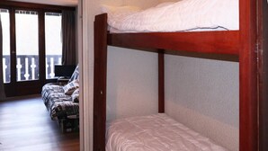 A bunk bed room with a wooden frame, a lower bunk with a white mattress, and a higher bunk with a white mattress. There is a window with a view of a balcony.