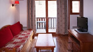A room with a wooden sofa, a wooden table, a television on a wooden stand, and a sliding glass door leading to an outdoor area.