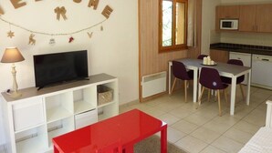 A compact living space with a TV, a red table, a dining area with chairs, a kitchen with cabinets, and a window with curtains. - N°101 Balcon des Airelles (LES ORRES 1800)