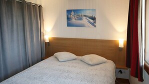 Un lit recouvert d’une couverture blanche à texture, avec deux oreillers, une tête de lit en bois et un tableau représentant un paysage enneigé accroché au mur.