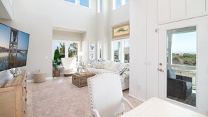 A spacious living room with a high ceiling, large windows, a white sofa, a dining table, and a view of the outdoors. - Hobbs Realty - “Carolina Pearl” | New! Inlet Views & Private Heatable Pool (Holden Beach)