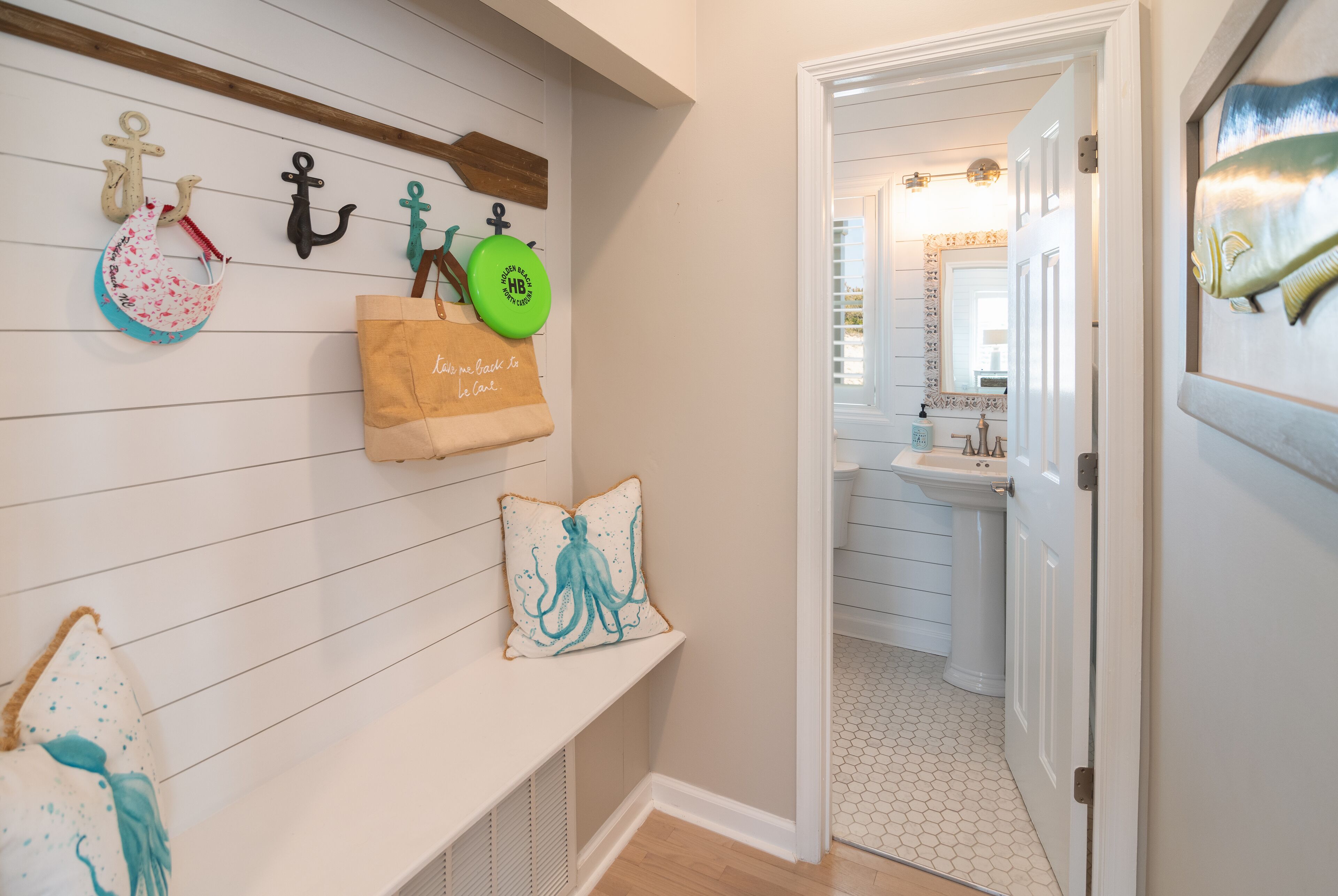 A hallway with a white bench, a wall-mounted shelf with decorative items, and a door leading to a bathroom with a sink and mirror.