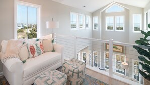 A spacious living room with a white couch, ottomans, and a view of the ocean through large windows. - Hobbs Realty - “Carolina Pearl” | New! Inlet Views & Private Heatable Pool (Holden Beach)
