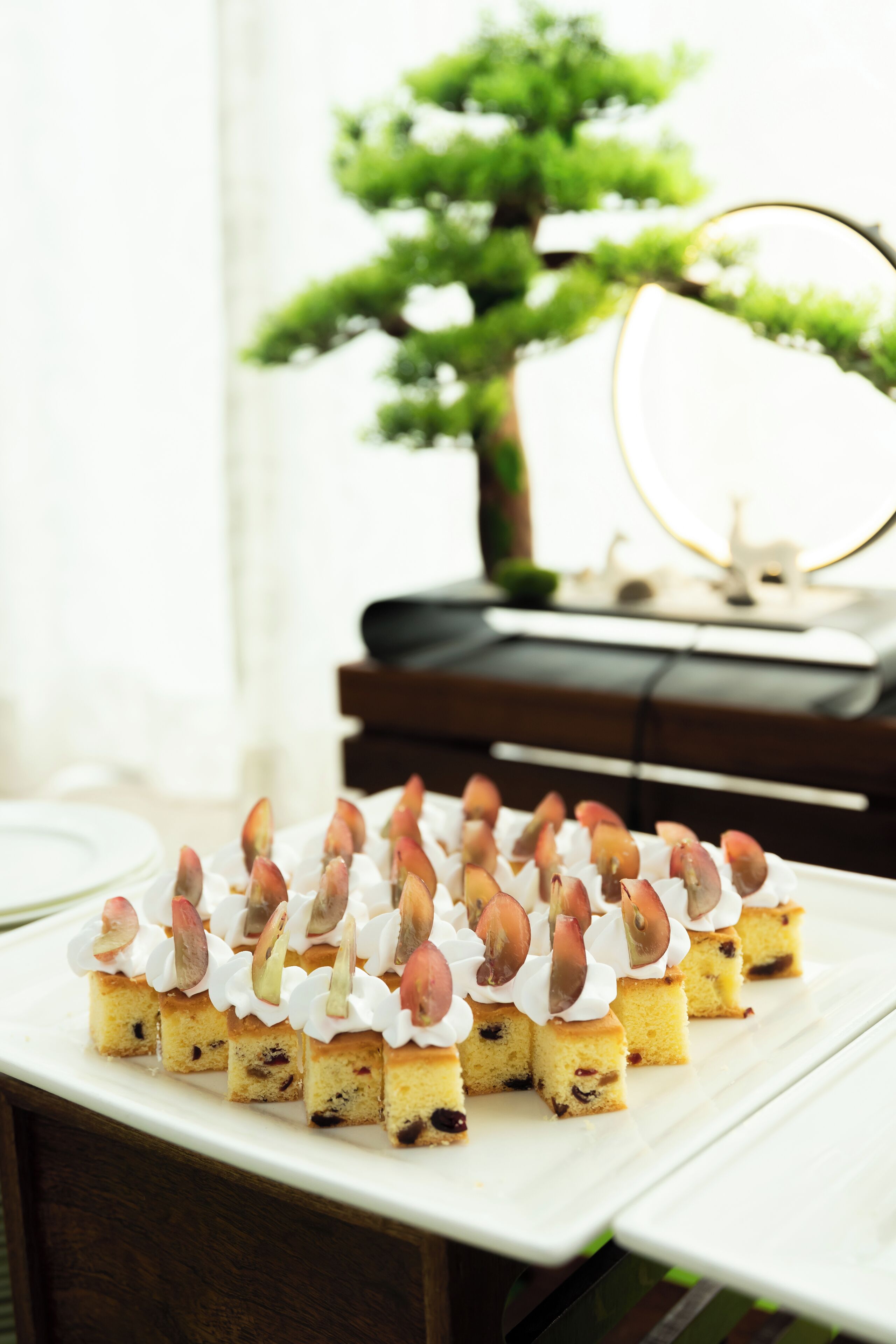 A dessert tray with small square cakes topped with whipped cream and almonds, placed on a white table in a room with a piano and a green plant in the background.