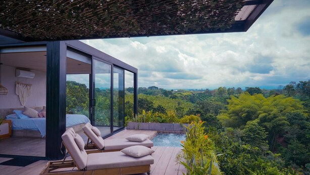 A modern bedroom with a large window, a wooden deck with lounge chairs, and a view of a lush landscape. - Amanatu Ecolodge & Spa  (Quimbaya)