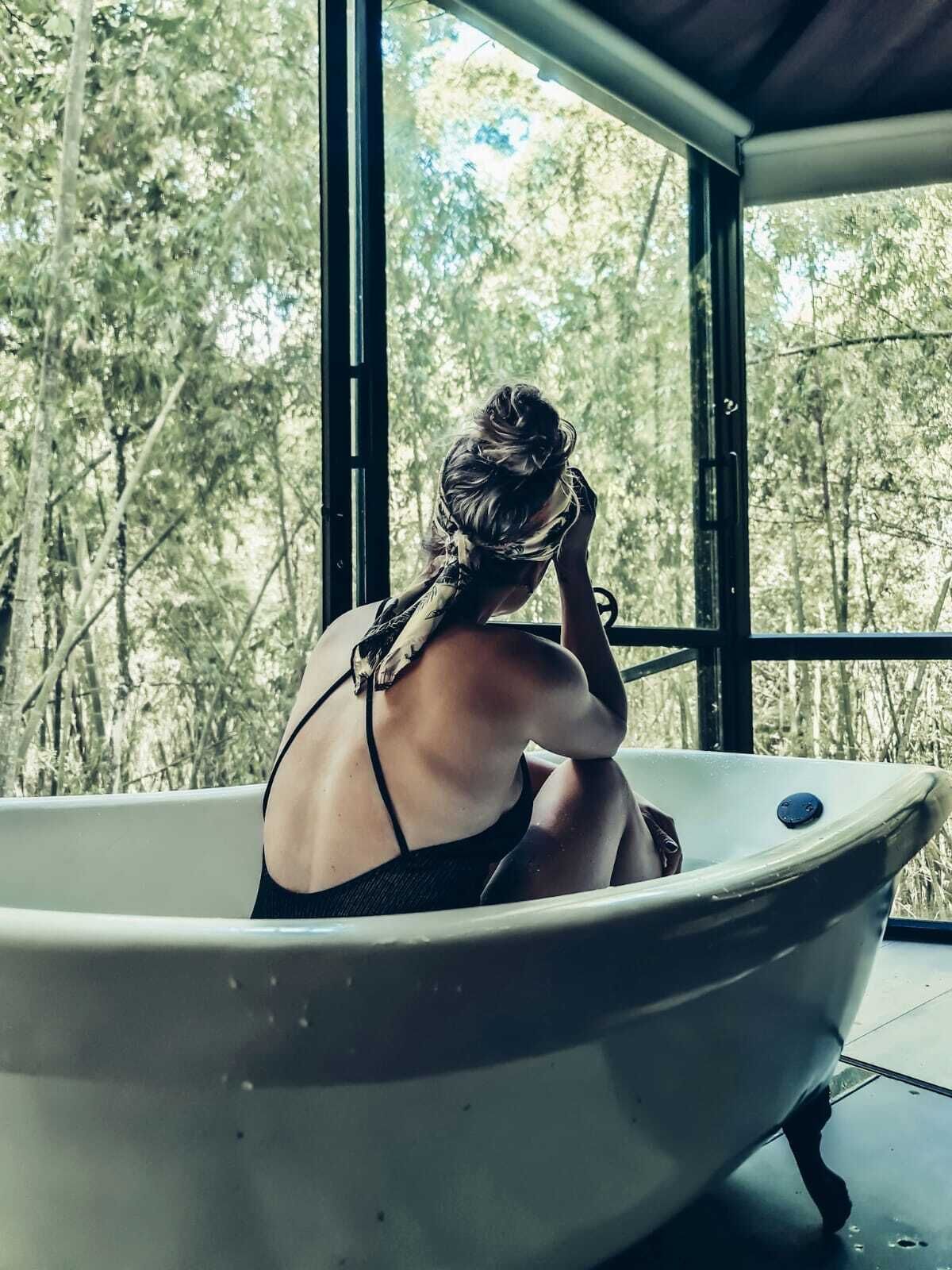 Una persona sentada en una bañera con vistas a árboles a través de amplios ventanales.