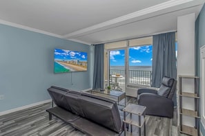 A modern living room with a large TV, a balcony with a view of the beach, and a seating area with a table.