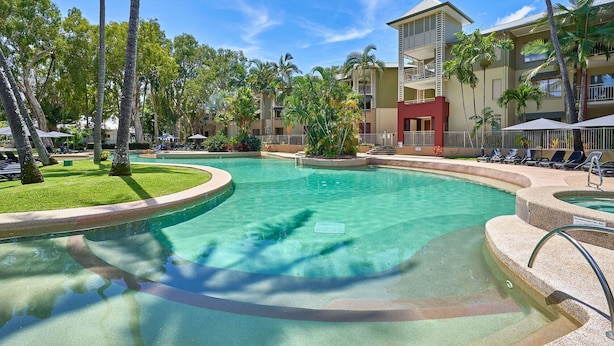 Una piscina de resort rodeada de palmeras y sillas de descanso, con un edificio que tiene balcones y un puesto de socorristas.