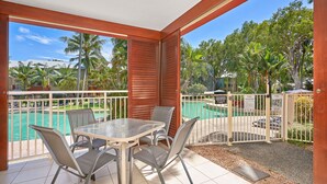 Un espacio al aire libre techado con piscina, sillas y vistas a una zona residencial con palmeras.