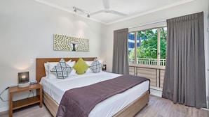 Un dormitorio con cama, mesita de noche, ventilador de techo, ventana con cortinas y vistas al exterior con vegetación.