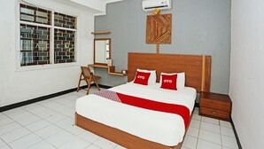 A bedroom with a wooden bed, two red pillows, a white bedspread, a wooden nightstand, a small wooden table, a chair, a mirror, and a window with a brick pattern. - OYO 92088 171 Inn (Bogor)