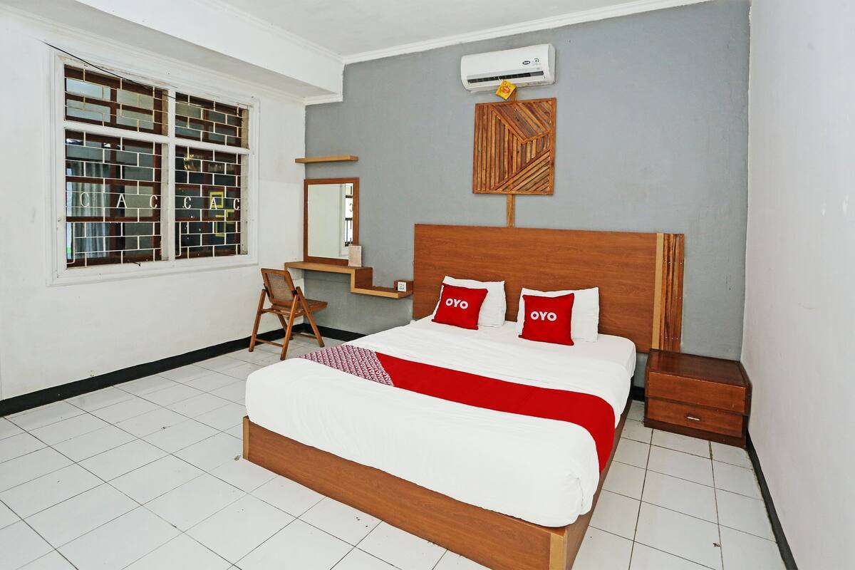 A bedroom with a wooden bed, two red pillows, a white bedspread, a wooden nightstand, a small wooden table, a chair, a mirror, and a window with a brick pattern.