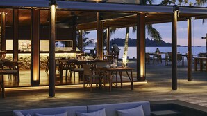 An outdoor dining area with wooden tables and chairs, a covered seating area with cushions, and a view of the sea. - The Retreat Koh Chang (Ko Chang)