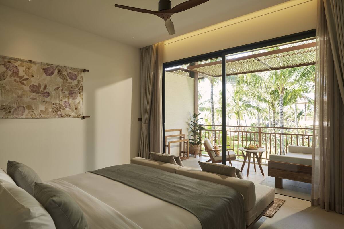 A bedroom with a large bed, a ceiling fan, and a view of the outdoor balcony with a table and chairs.