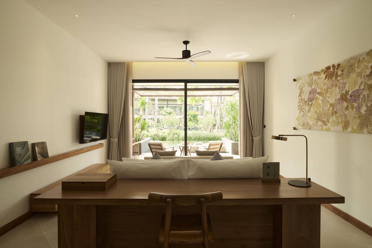A modern room with a wooden desk, a ceiling fan, a large window with curtains, a small table with chairs, and a wall-mounted TV.