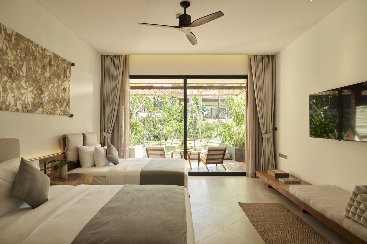 A modern hotel room with two beds, a ceiling fan, a large window with a view of greenery, and a flat-screen TV.
