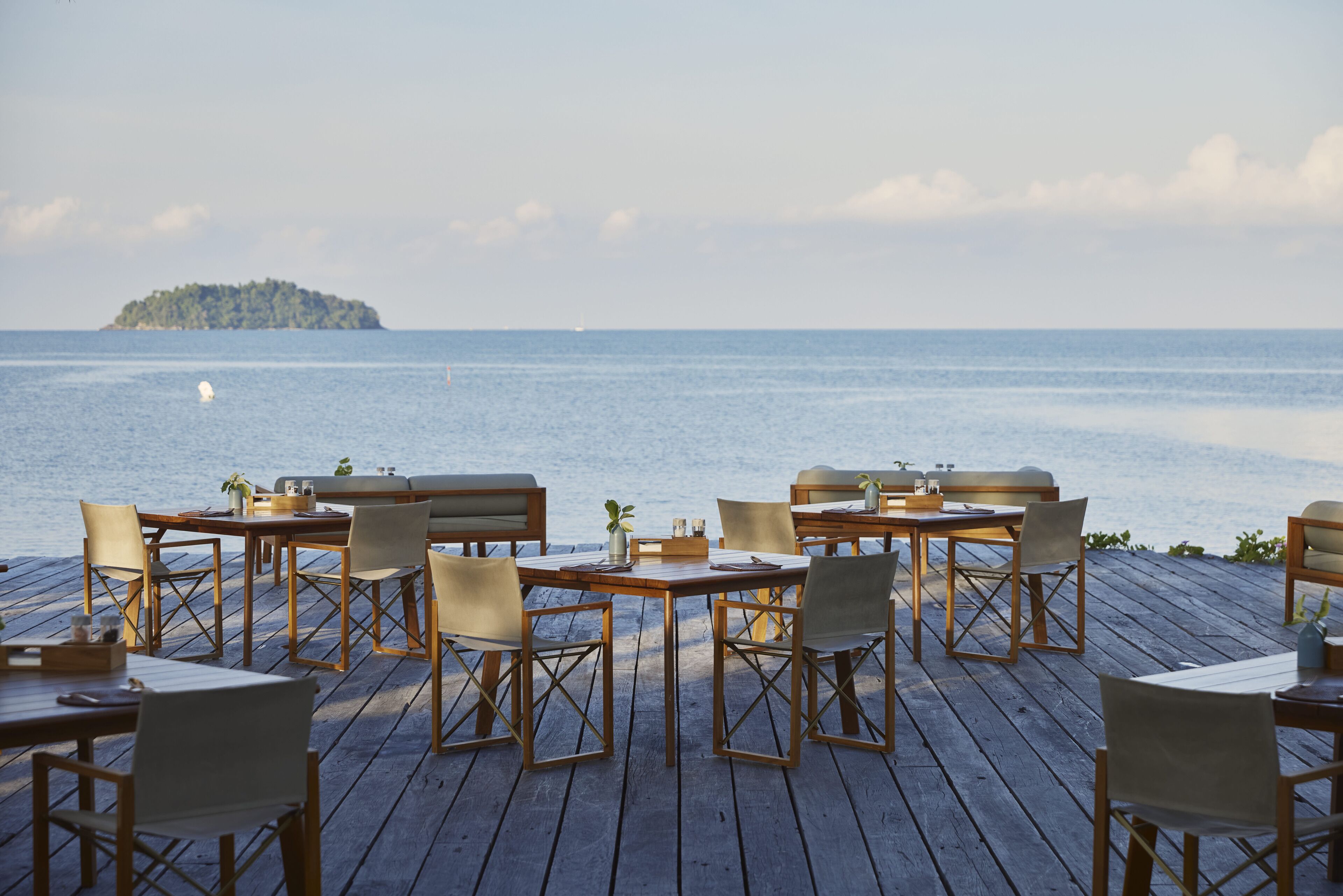 A wooden deck with tables and chairs facing a calm sea and a distant island.