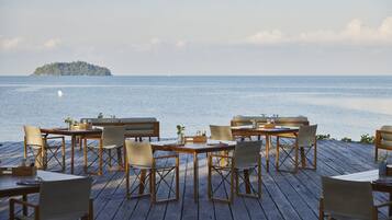 A wooden deck with tables and chairs facing a calm sea and a distant island.