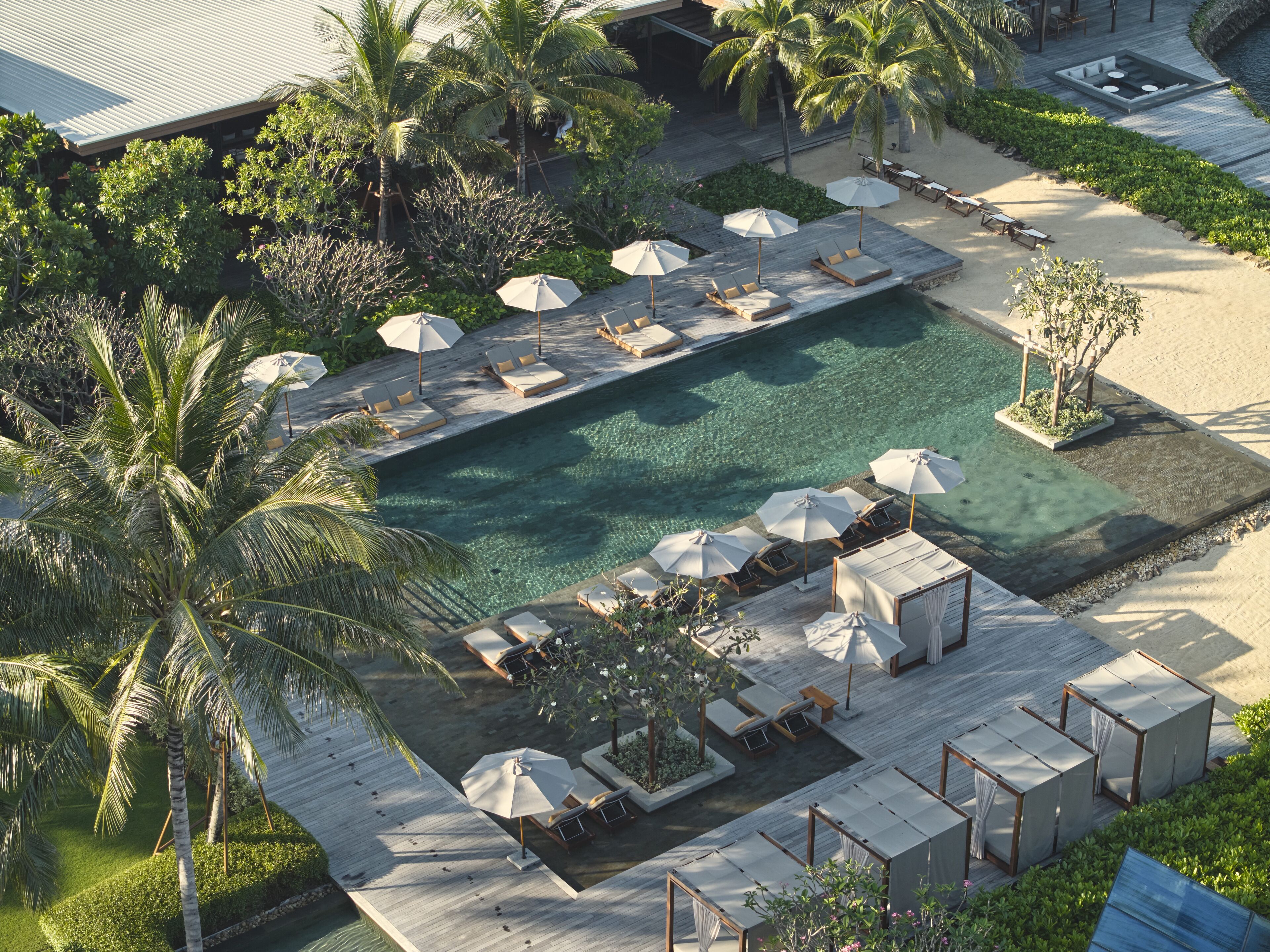 An aerial view of a resort with a swimming pool, lounge areas with umbrellas, and palm trees.