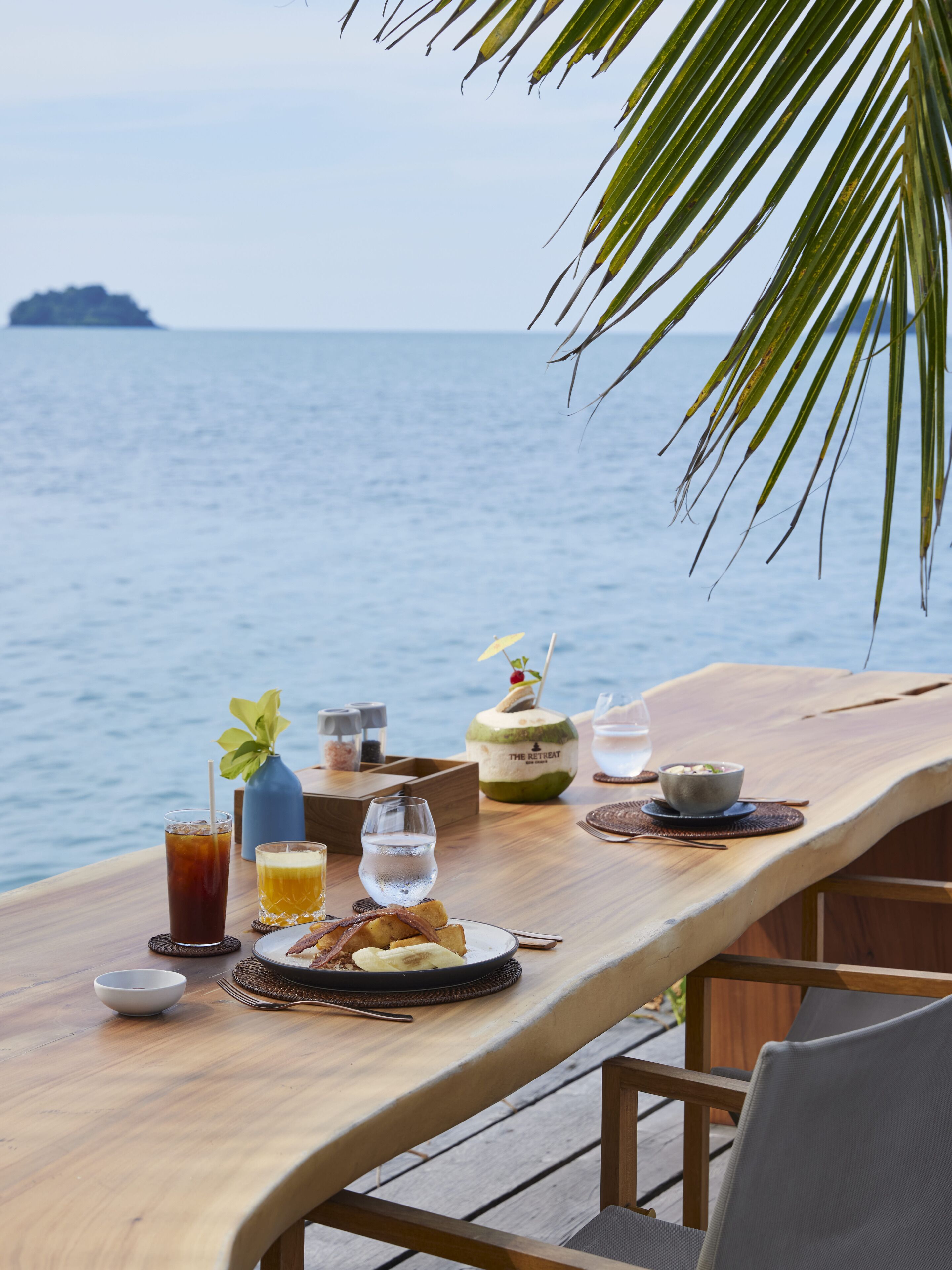 A wooden table set with a glass of iced tea, a glass of water, a bowl of soup, and a plate of food, overlooking a body of water with a palm tree in the foreground.