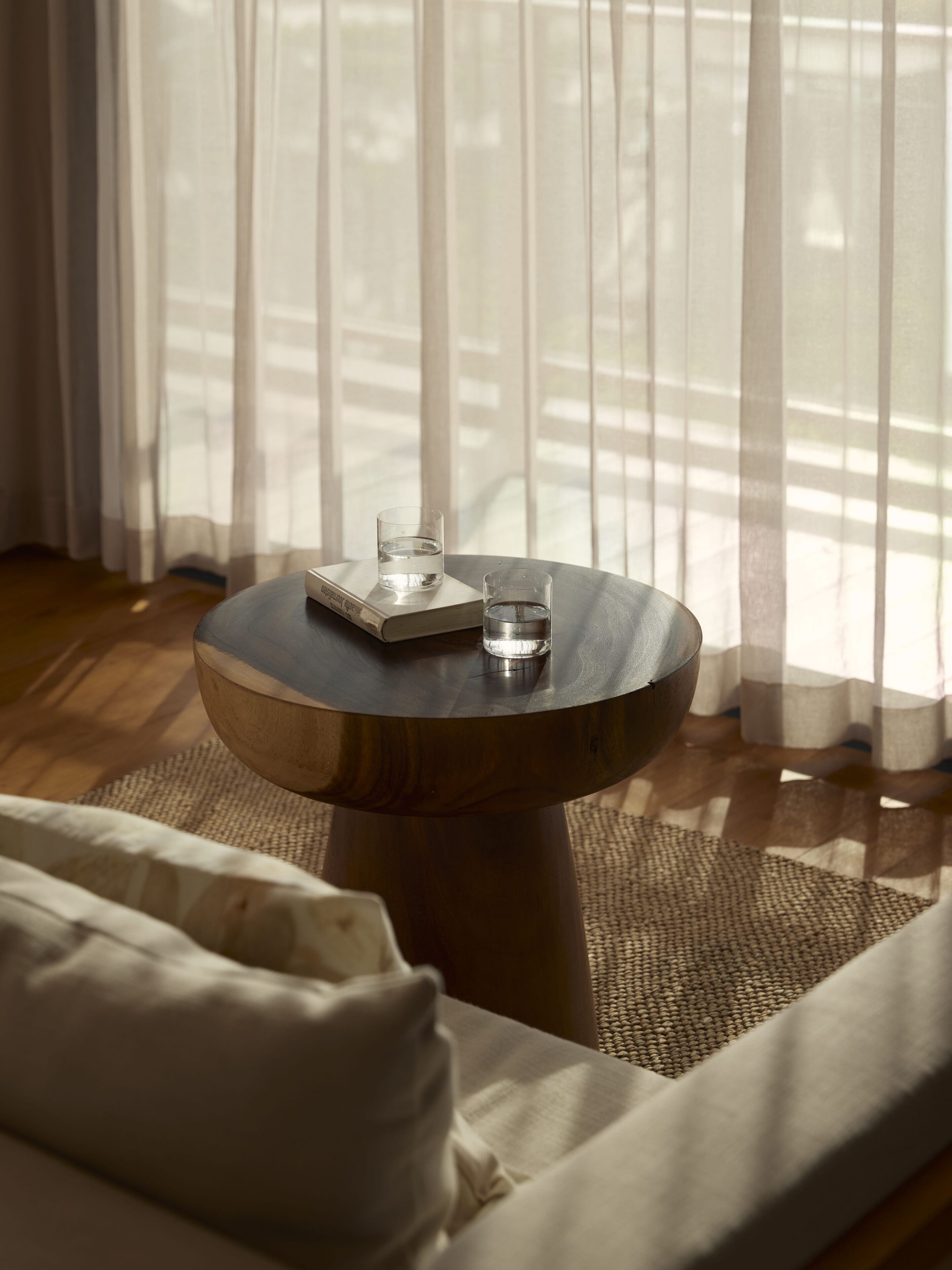 Una mesa redonda de madera con dos vasos de agua y una bandeja, sobre una alfombra tejida en una habitación con un sofá y cortinas translúcidas.