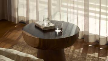 Una mesa redonda de madera con dos vasos de agua y una bandeja, sobre una alfombra tejida en una habitación con un sofá y cortinas translúcidas.