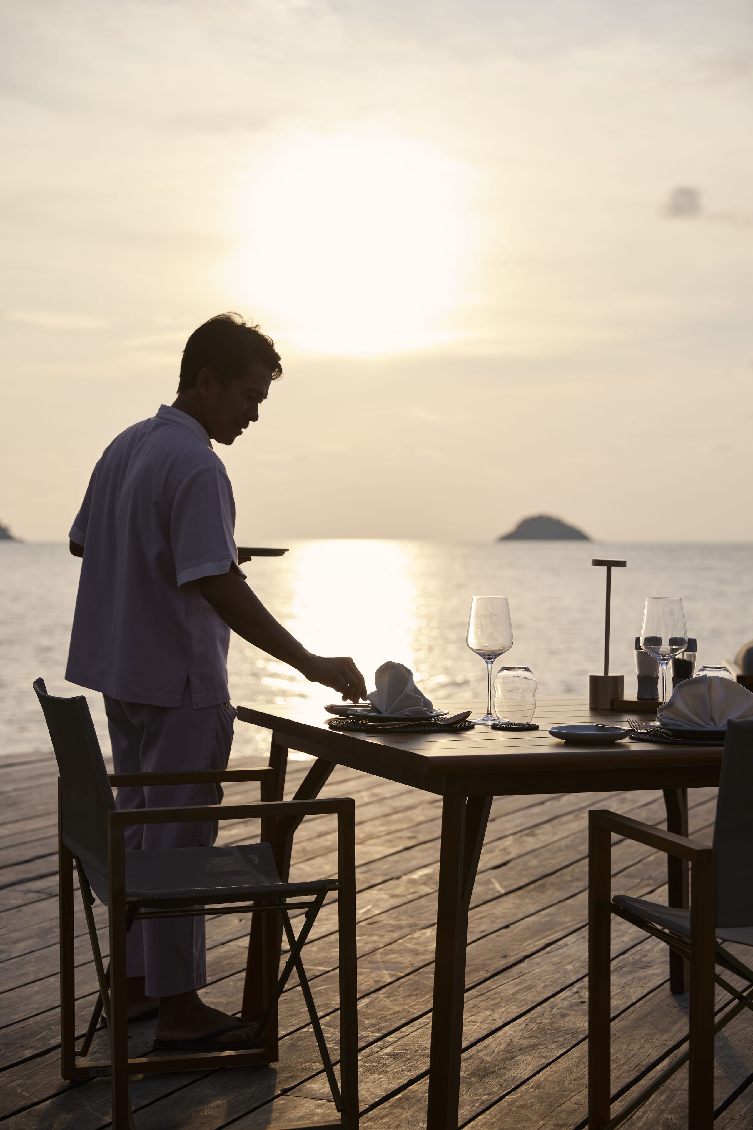 A wooden deck with a table set for two, overlooking a calm sea with a distant island.