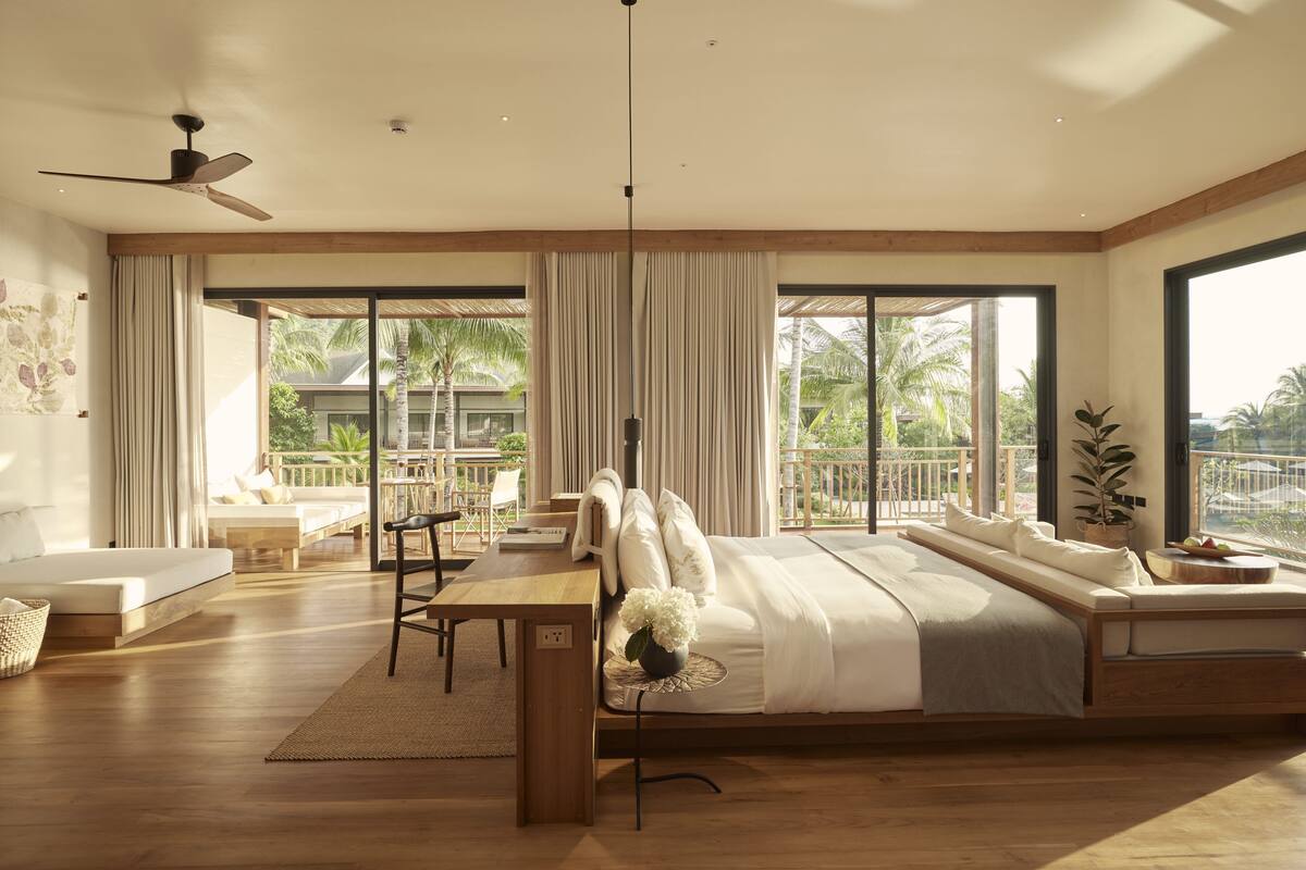 A spacious bedroom with a large bed, a wooden desk, a dining area, and a balcony with a view of palm trees.