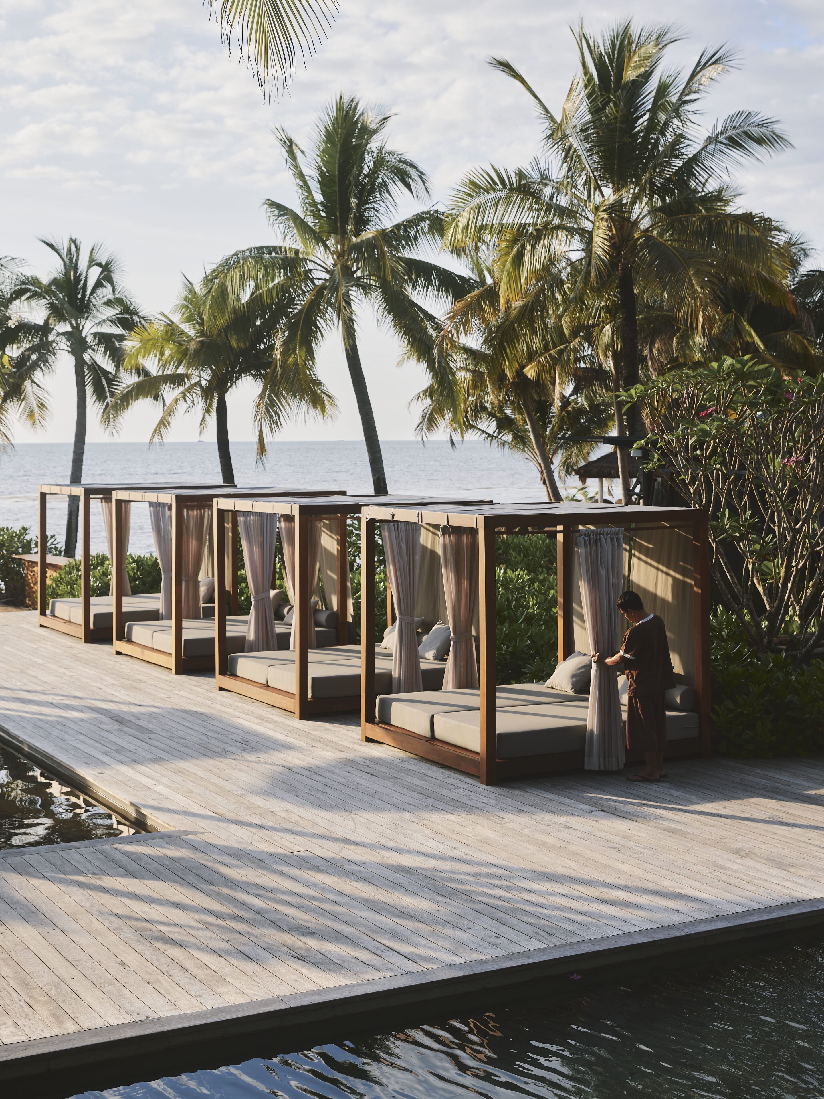 A wooden deck with four canopy-style beds overlooking a body of water, surrounded by palm trees.