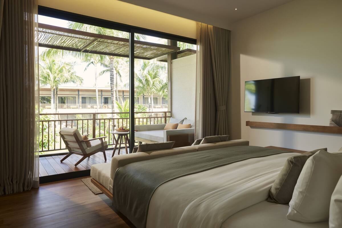 A hotel room with a large bed, a TV mounted on the wall, a balcony with a seating area, and a view of palm trees.