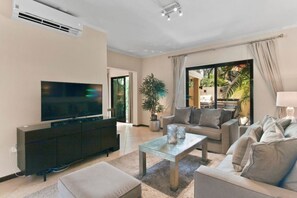A modern living room with a flat-screen TV, a dark entertainment unit, a glass coffee table, and a beige sofa.