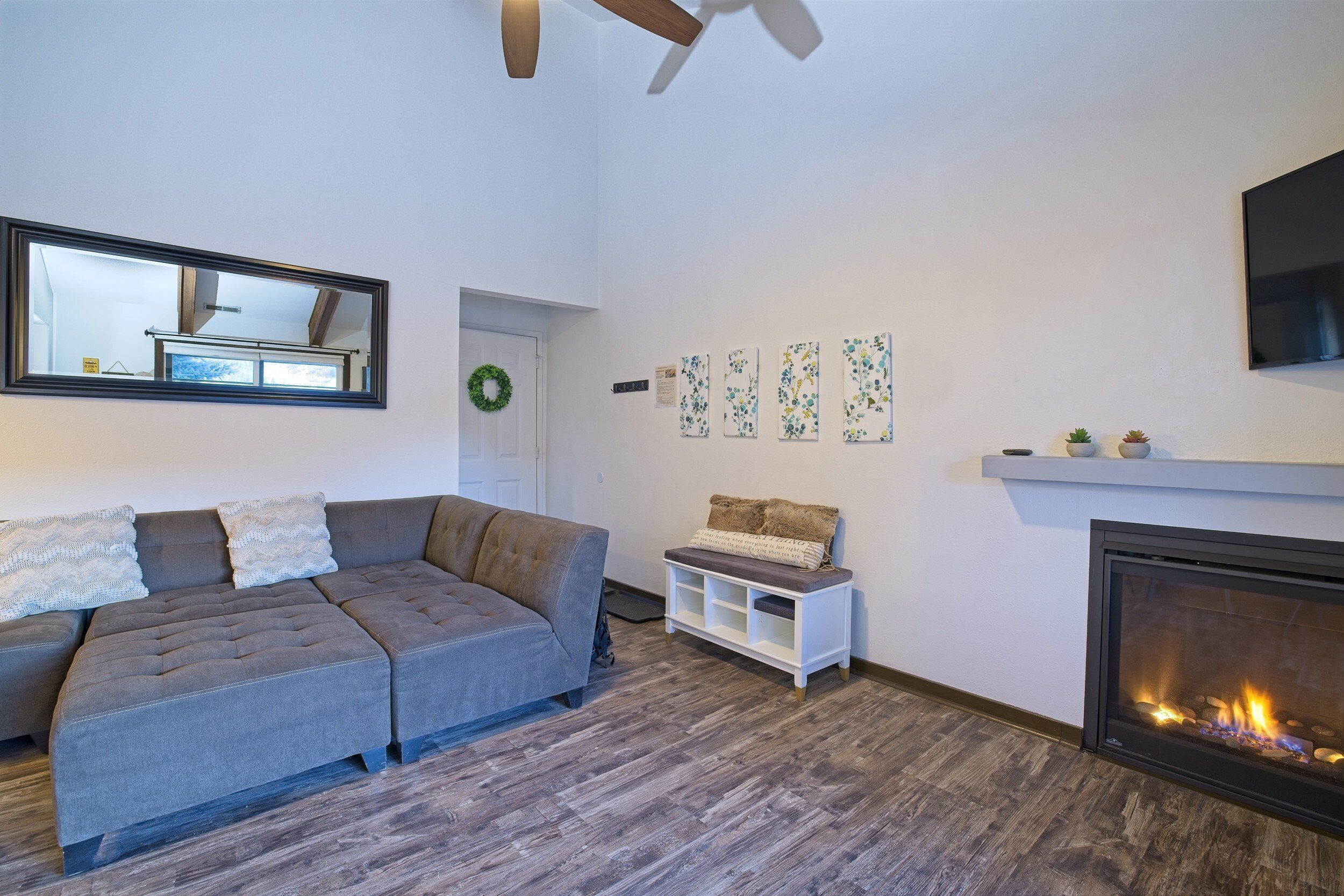 Ein Wohnzimmer mit grauem Ecksofa, einem weißen Beistelltisch, einem Kamin und einem Wand-TV.
