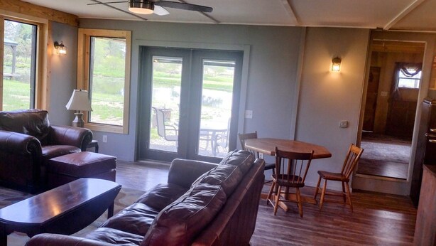 A living room with a sofa, coffee table, and dining table with chairs. There are large windows and a view of the outdoors. - Minutes from Pymatuning, Cabin by the Pond (Williamsfield)