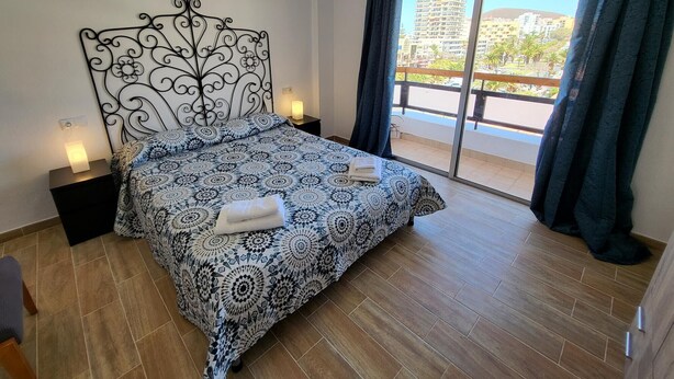 A bedroom with a patterned bedspread, a decorative headboard, a nightstand with a lamp, and a view of a balcony with a cityscape. - Jardines Canarios 3 (Los Cristianos)