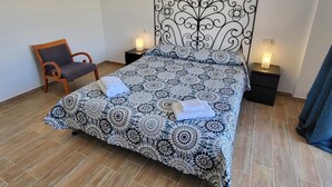 A neatly made bed with a patterned bedspread, two bedside tables with lamps, a wooden chair, and a blue curtain. - Jardines Canarios 3 (Los Cristianos)