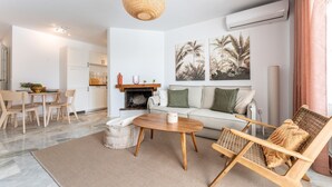 En moderne stue med sofa, sofabord og fletstole. Der er en pejs, et spiseområde med bord og stole, og et køkken i baggrunden.