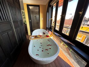 A bathroom with a freestanding bathtub, a shower, and a window with a view of buildings. - Hoianan Boutique Hotel (Da Nang)